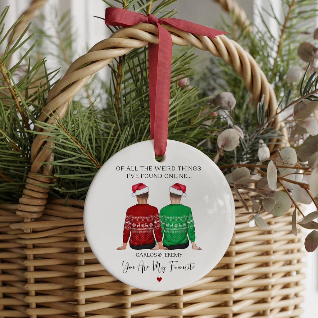 A round white ceramic Christmas ornament hanging from a red ribbon bow in a wicker basket, decorated with an illustration of two people in red and green sweaters and a humorous caption.
