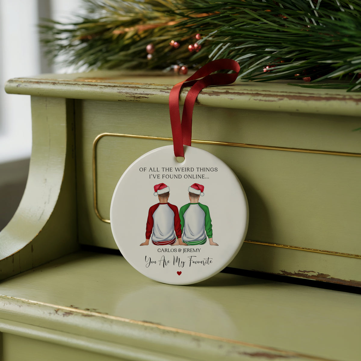 A round ceramic Christmas ornament with a red ribbon hanger, featuring a printed illustration of two boys in Santa hats and personalized text, displayed on a distressed sage green wooden cabinet with pine branches in the background.