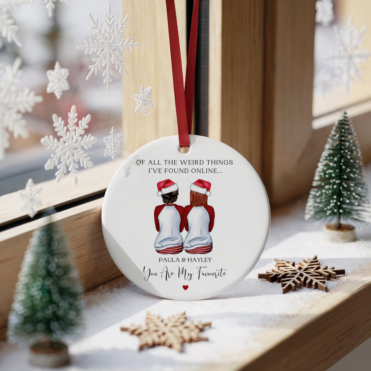 A personalized round white Christmas ornament hanging on a wooden window sill, decorated with a red ribbon. The ornament features a printed illustration of a couple in red sweaters and Santa hats, with the text 'OF ALL THE WEIRD THINGS I'VE FOUND ONLINE.