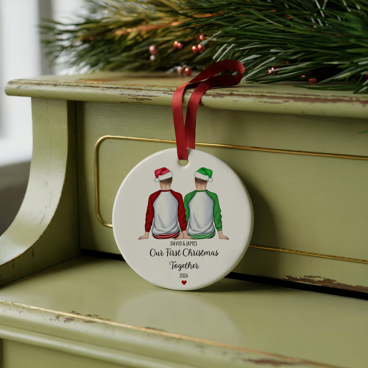A round white Christmas ornament with a red ribbon hanging from the top, featuring an illustration of two people from behind wearing red and green shirts and Santa hats, with the text 'SMITH & WATSON Our True Christmas Together 2026'. The ornament is displayed on a distressed sage green wooden cabinet with pine branches in the background.
