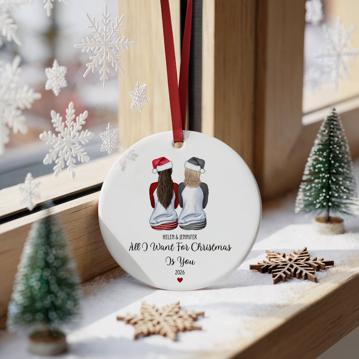 A round white Christmas ornament hanging on a wooden windowsill, decorated with an illustration of two girls from behind wearing a red and black hat. The ornament has a red hanging ribbon and printed text reading 'BELLAD & JENNIFER All I Want For Christmas Is You 2026'.