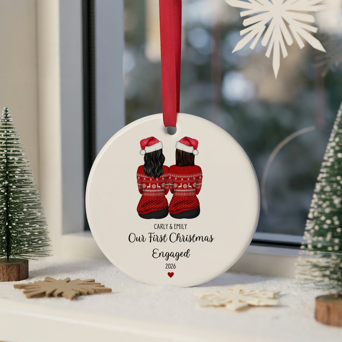 A personalized round white Christmas ornament featuring an illustration of a couple in red Santa hats and sweaters, hanging by a red ribbon. The ornament is printed with the text 'CARLY & EMILY Our First Christmas Engaged 2026'.