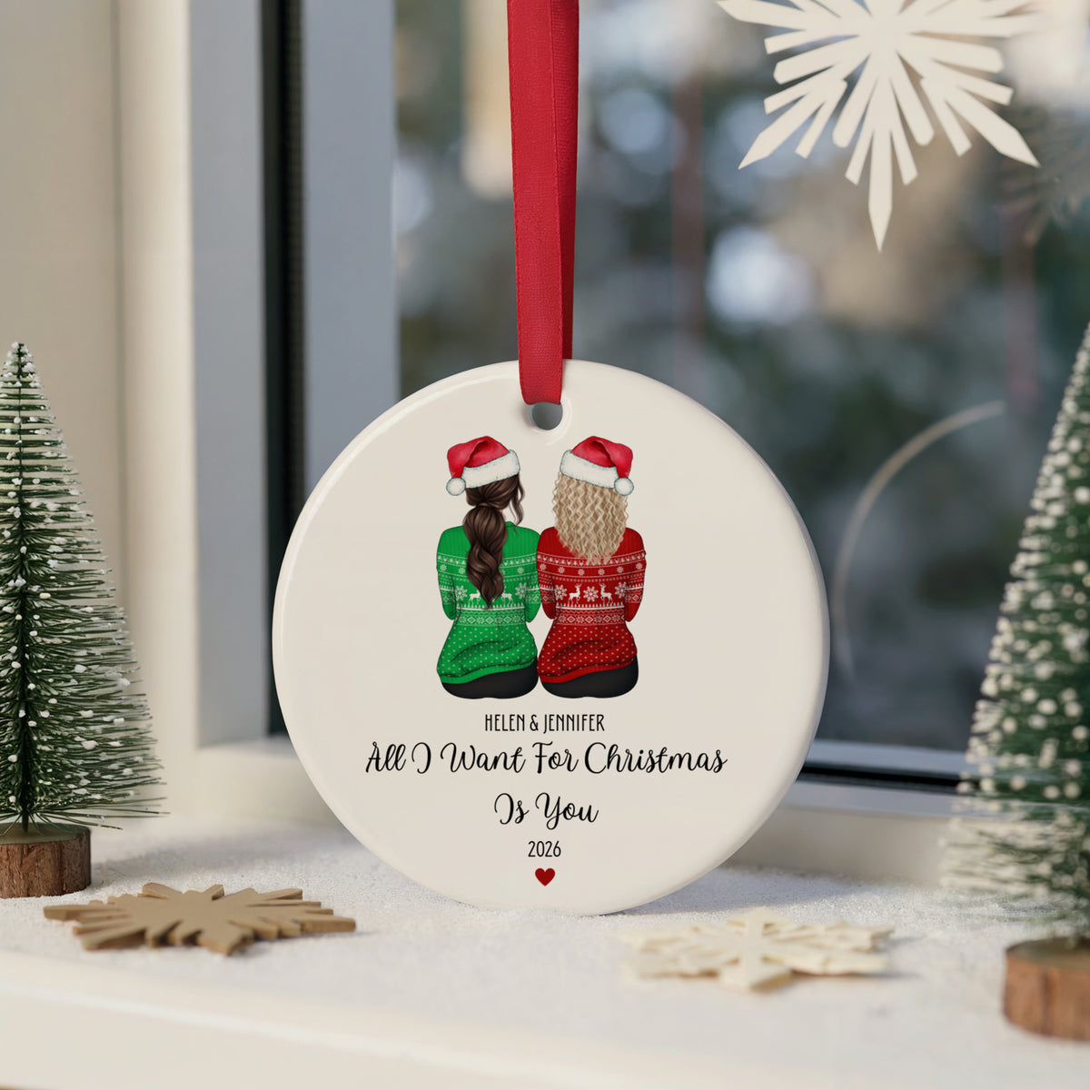 A round white Christmas ornament hanging from a red ribbon, featuring an illustration of two women in Santa hats and sweaters, with personalized text reading 'HELEN & JENNIFER All I Want For Christmas Is You 2026'. The ornament is displayed on a windowsill with small decorative trees and snowflake cutouts.