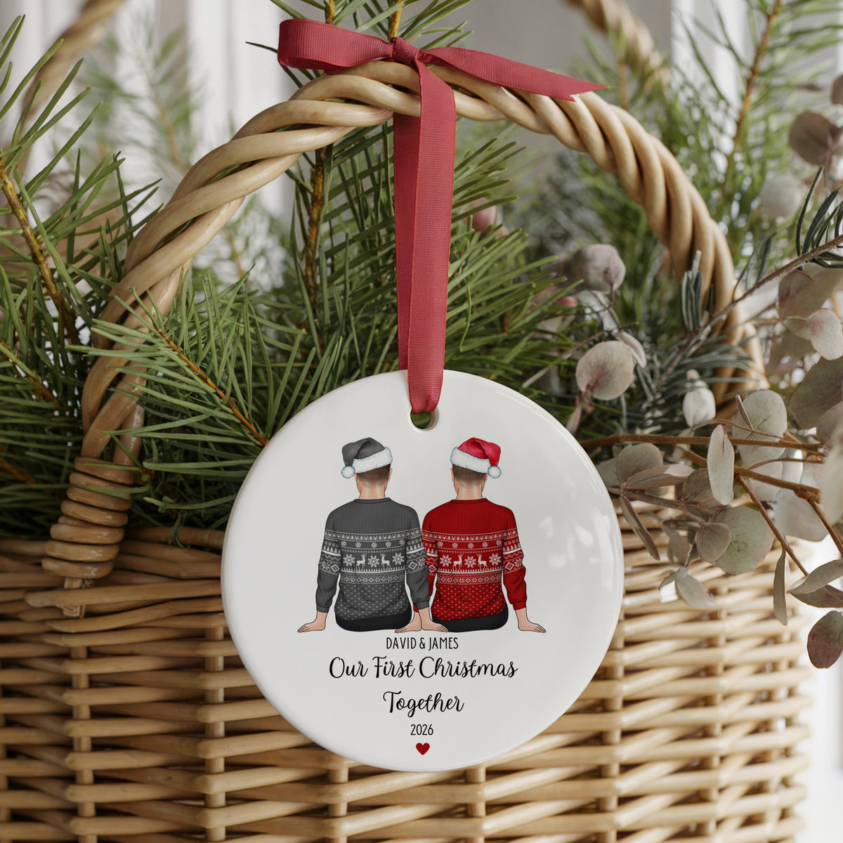 A round white ceramic Christmas ornament hanging from a red ribbon in a wicker basket with pine branches. The ornament features an illustration of two men seen from behind, one in a gray sweater and one in a red sweater, with the personalized text 'DAVID & RAMES Our First Christmas Together 2026'.