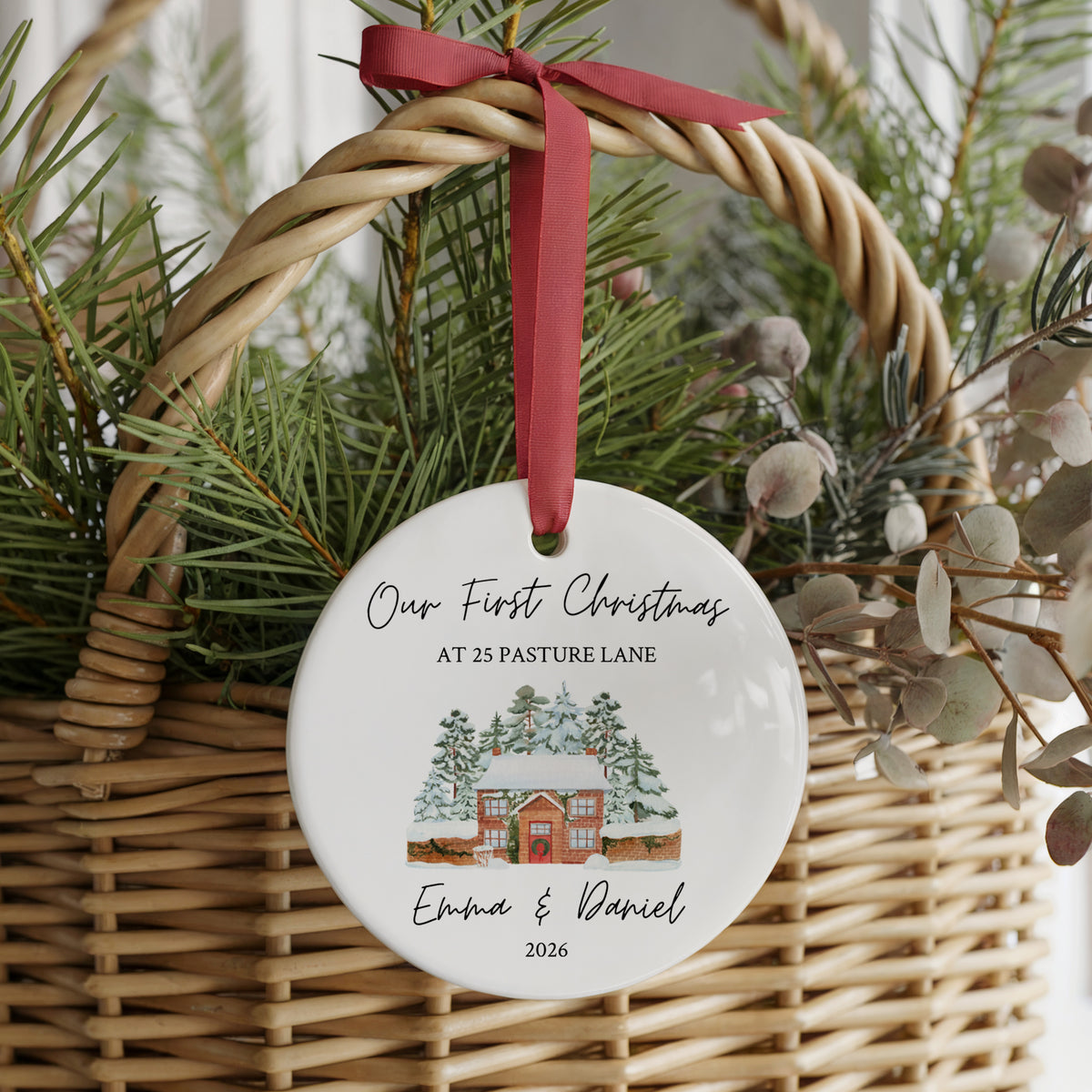 A round white ceramic Christmas ornament hanging from a red ribbon bow in a wicker basket filled with greenery. The ornament is personalized with the text 'Our First Christmas AT 25 PASTURE LANE Emma & Daniel 2026' above a small watercolor cabin illustration.