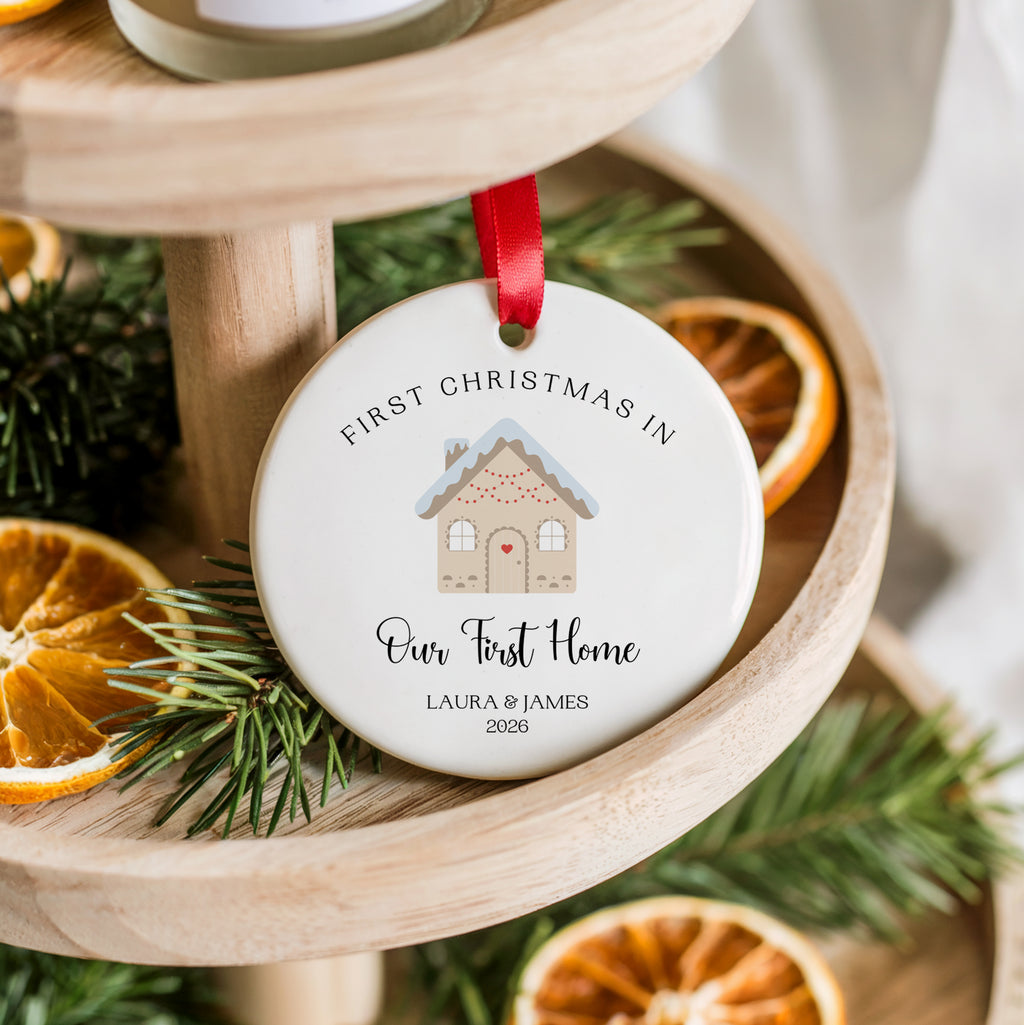 A round white ceramic Christmas ornament hanging from a red ribbon, printed with a small gingerbread house illustration and the text 'FIRST CHRISTMAS IN Our First Home LAURA & JAMES 2026.' The ornament is styled on a wooden tiered stand with pine branches and dried orange slices.