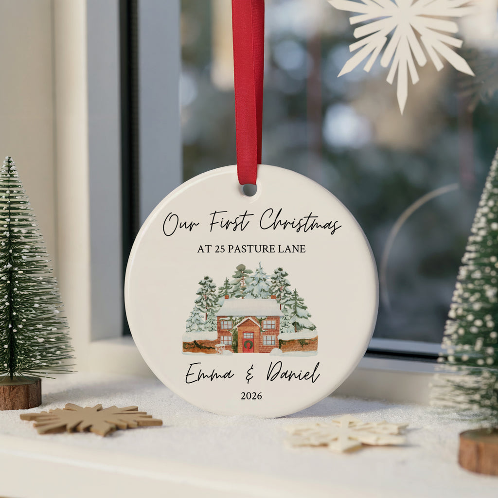 A personalized round ceramic Christmas ornament hanging on a windowsill. The ornament is white with a red ribbon, printed with the text 'Our First Christmas AT 25 PASTURE LANE' and 'Emma & Daniel 2026' around an illustration of a snowy cabin.