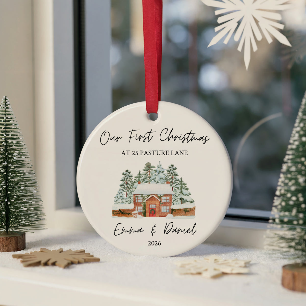 A personalized round ceramic Christmas ornament hanging on a windowsill. The ornament is white with a red ribbon, printed with the text 'Our First Christmas AT 25 PASTURE LANE' and 'Emma & Daniel 2026' around an illustration of a snowy cabin.