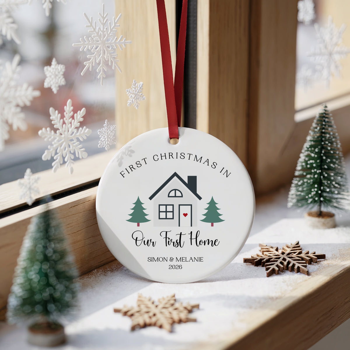 A round white personalized Christmas ornament hanging from a red ribbon, printed with a house illustration and the text 'FIRST CHRISTMAS IN Our First Home SIMON & MELANIE 2025'. The ornament is styled on a windowsill with small green trees, wooden snowflake decorations, and a soft window background.