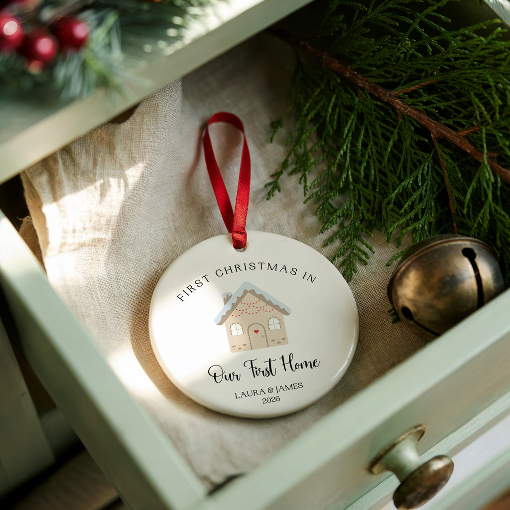 A personalized round Christmas ornament with a red ribbon, displayed in a light green drawer surrounded by pine branches, a small brass bell, and a beige fabric lining. The ornament is printed with 'FIRST CHRISTMAS IN Our First Home' and a small house illustration.