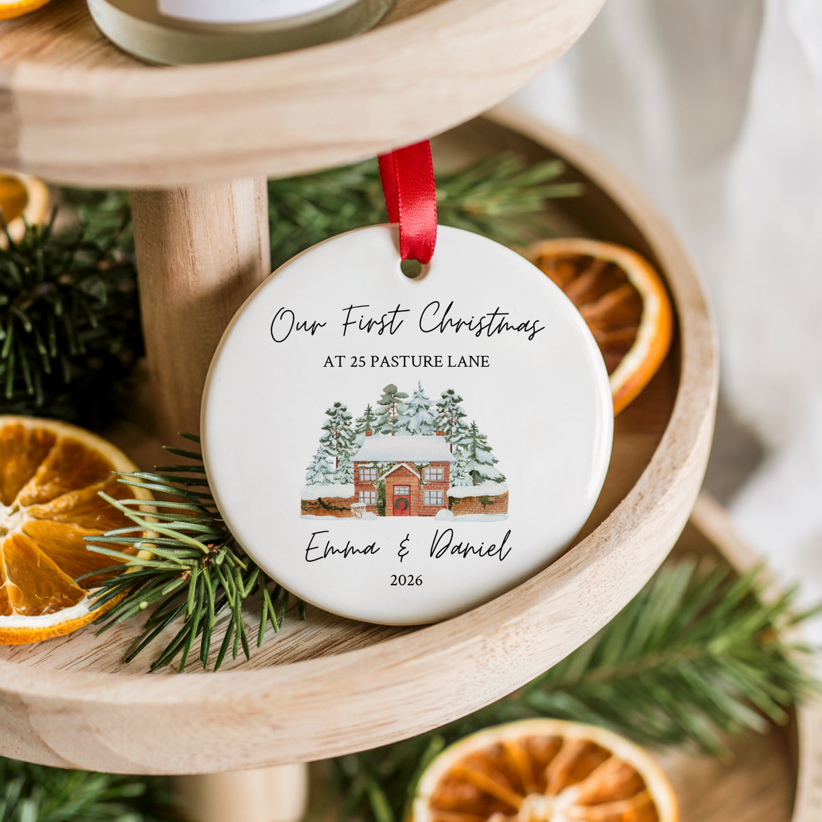 A round white ceramic Christmas ornament with a red ribbon, featuring a personalized winter cabin illustration and the text 'Our First Christmas AT 25 PASTURE LANE Emma & Daniel 2026', styled on a wooden tiered stand with pine branches and dried orange slices.