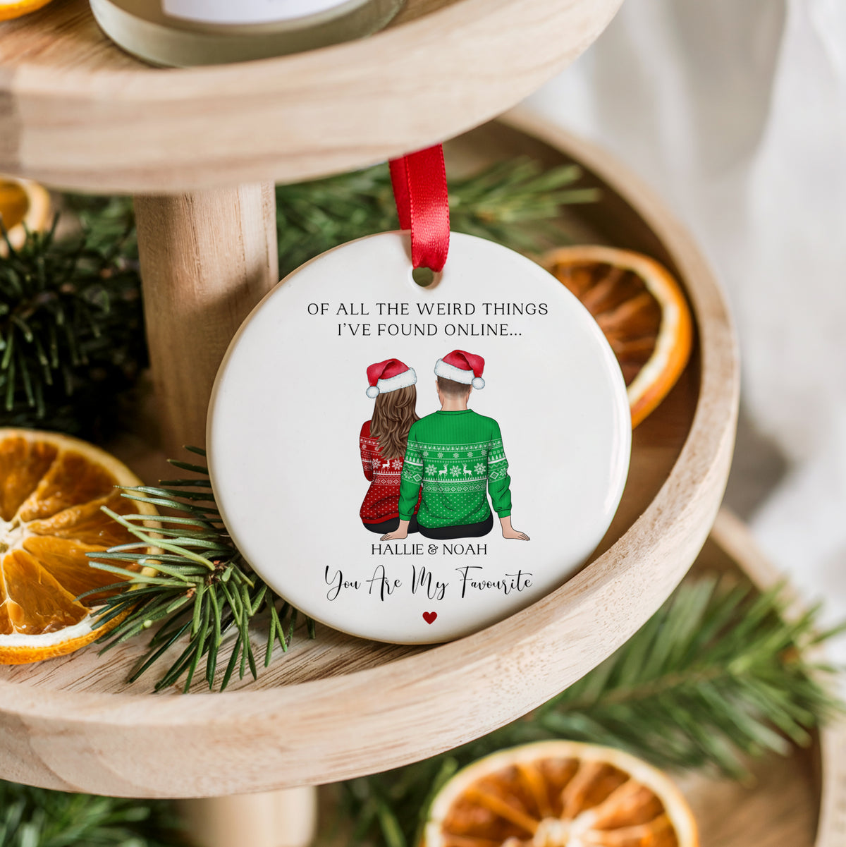 A round white ceramic Christmas ornament hanging from a red ribbon, featuring a printed illustration of a couple in Santa hats and the text 'OF ALL THE WEIRD THINGS I'VE FOUND ONLINE.