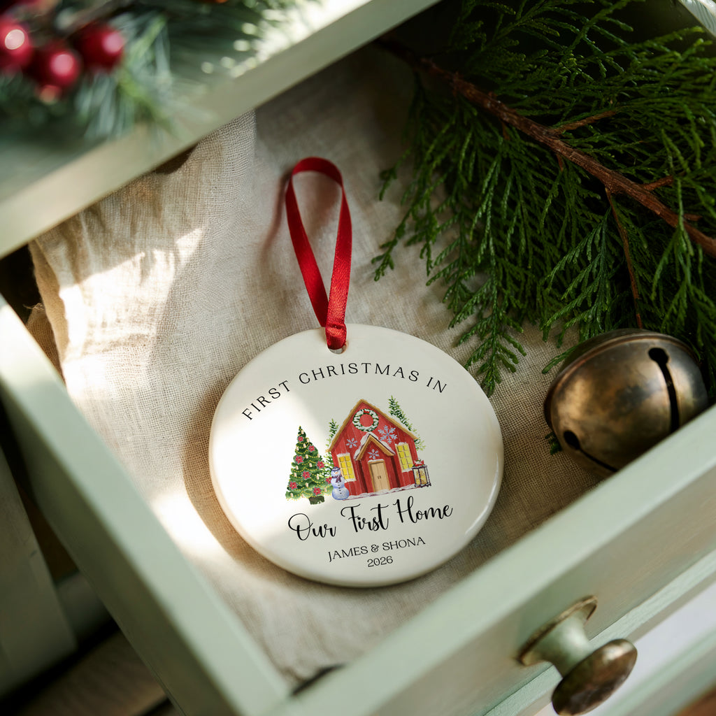 A round white personalized Christmas ornament with a red ribbon, featuring a printed illustration of a red house and Christmas tree, resting in a light green drawer surrounded by pine branches, a brass bell, and fabric.