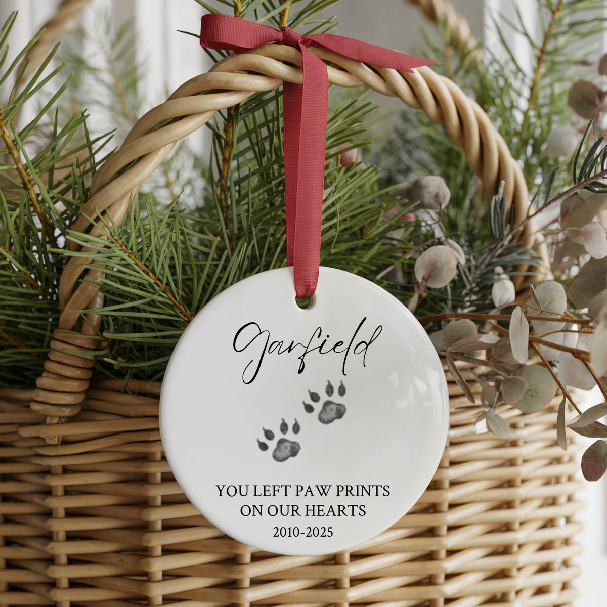A round white ceramic pet memorial ornament hanging from a red ribbon inside a wicker basket with evergreen branches. The ornament is personalized with the name 'Garfield' and paw prints, with the text 'YOU LEFT PAW PRINTS ON OUR HEARTS 2010-2025'.