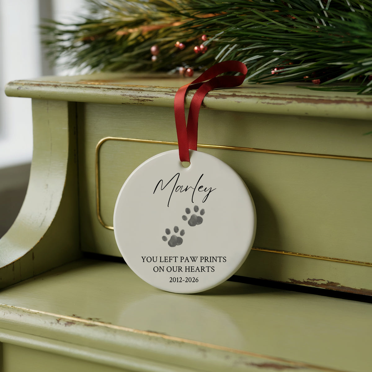 A round white pet memorial ornament hanging from a red ribbon on a distressed green cabinet, with pine branches and small red ornaments in the background. The ornament is printed with the name 'Marley', gray paw prints, and the text 'YOU LEFT PAW PRINTS ON OUR HEARTS 2012-2026'.