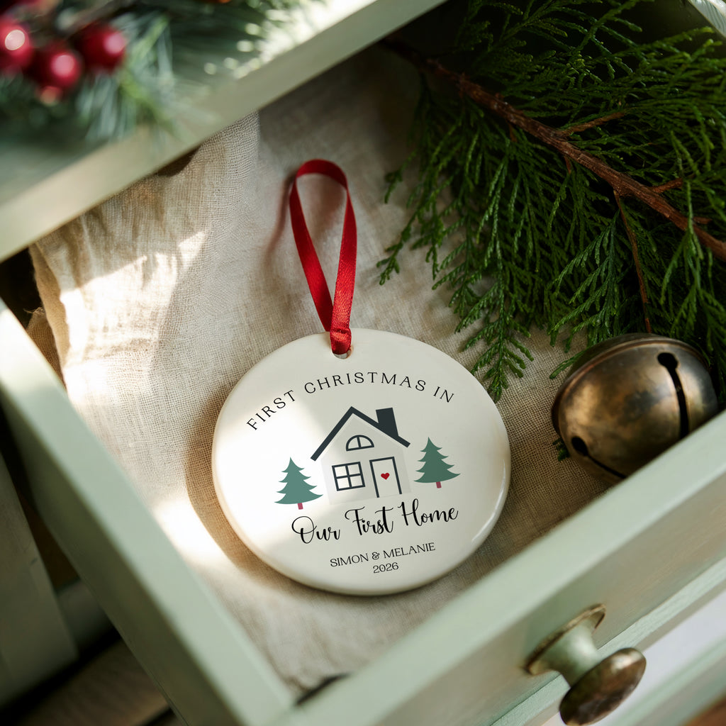 A round white Christmas ornament with a red ribbon, printed with 'FIRST CHRISTMAS IN Our First Home' and a small house and tree illustration, resting in an open drawer with pine branches and a brass bell nearby.